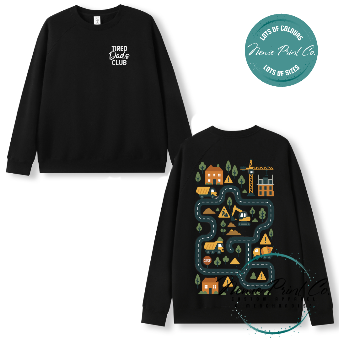 Tired Dads Club black t-shirt featuring front text and a vibrant construction-themed illustration on the back with houses, vehicles, and winding roads. Available in multiple t-shirt colors and various jumper options. Designed and printed by Newie Print Co.