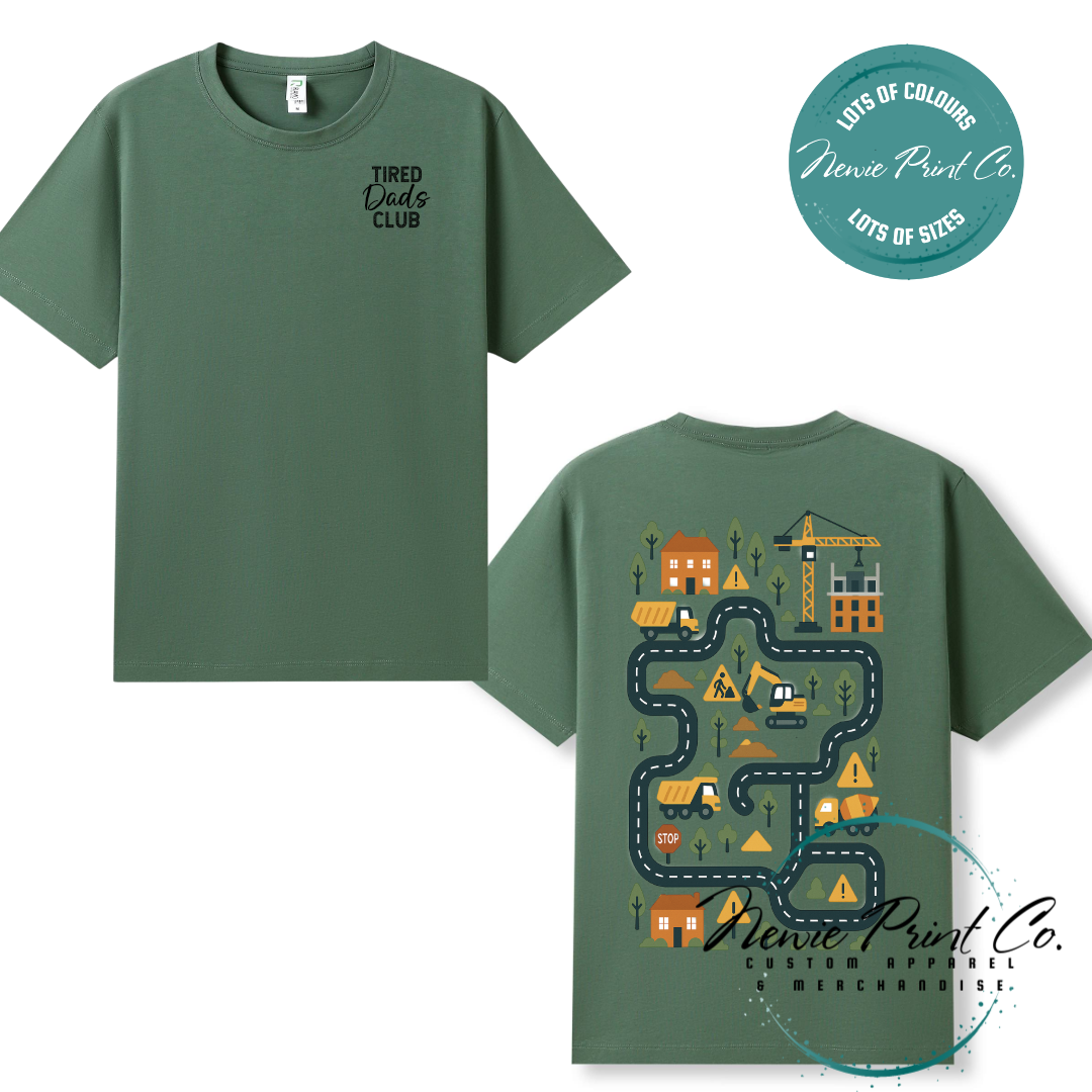 Tired Dads Club black t-shirt featuring front text and a vibrant construction-themed illustration on the back with houses, vehicles, and winding roads. Available in multiple t-shirt colors and various jumper options. Designed and printed by Newie Print Co.