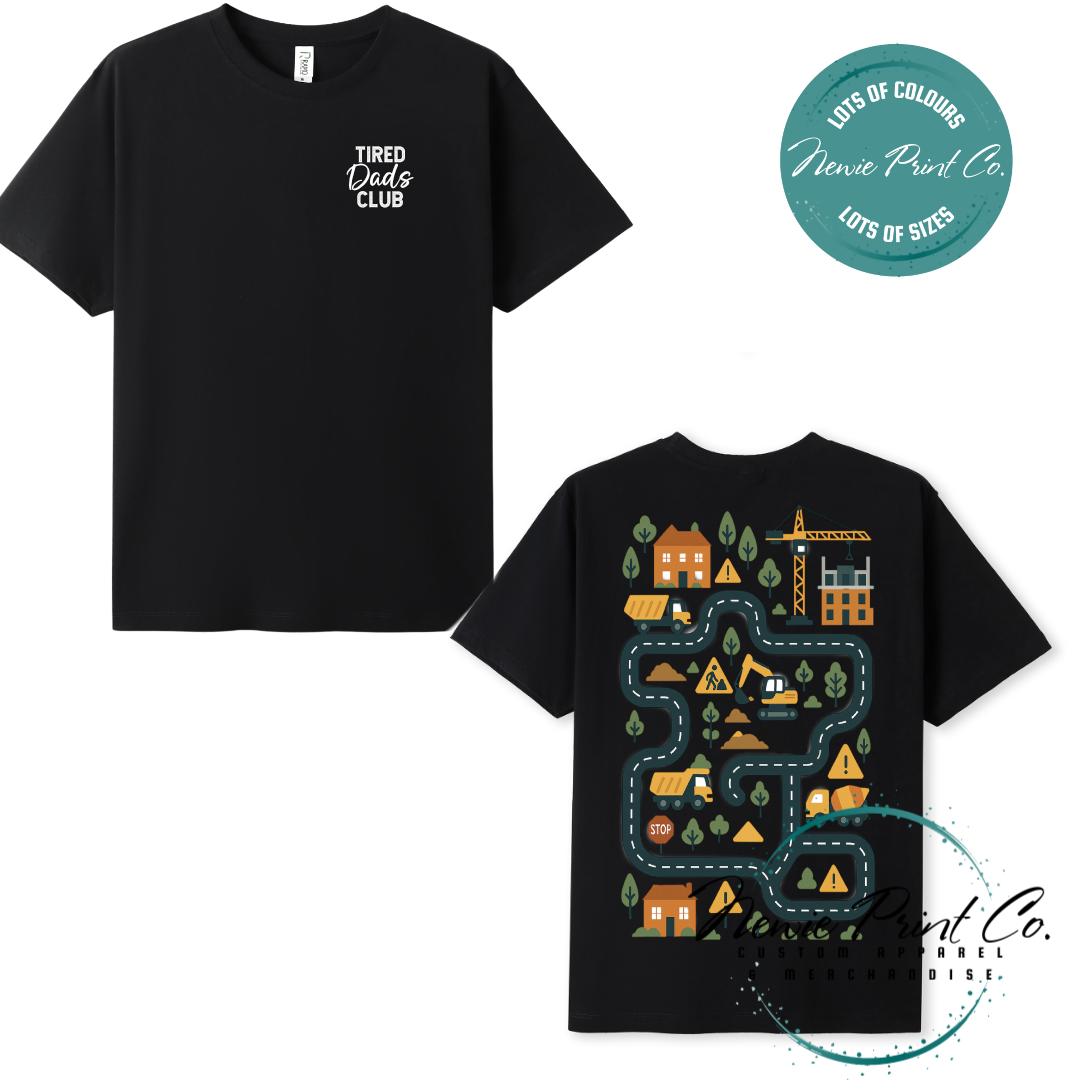 Tired Dads Club black t-shirt featuring front text and a vibrant construction-themed illustration on the back with houses, vehicles, and winding roads. Available in multiple t-shirt colors and various jumper options. Designed and printed by Newie Print Co.