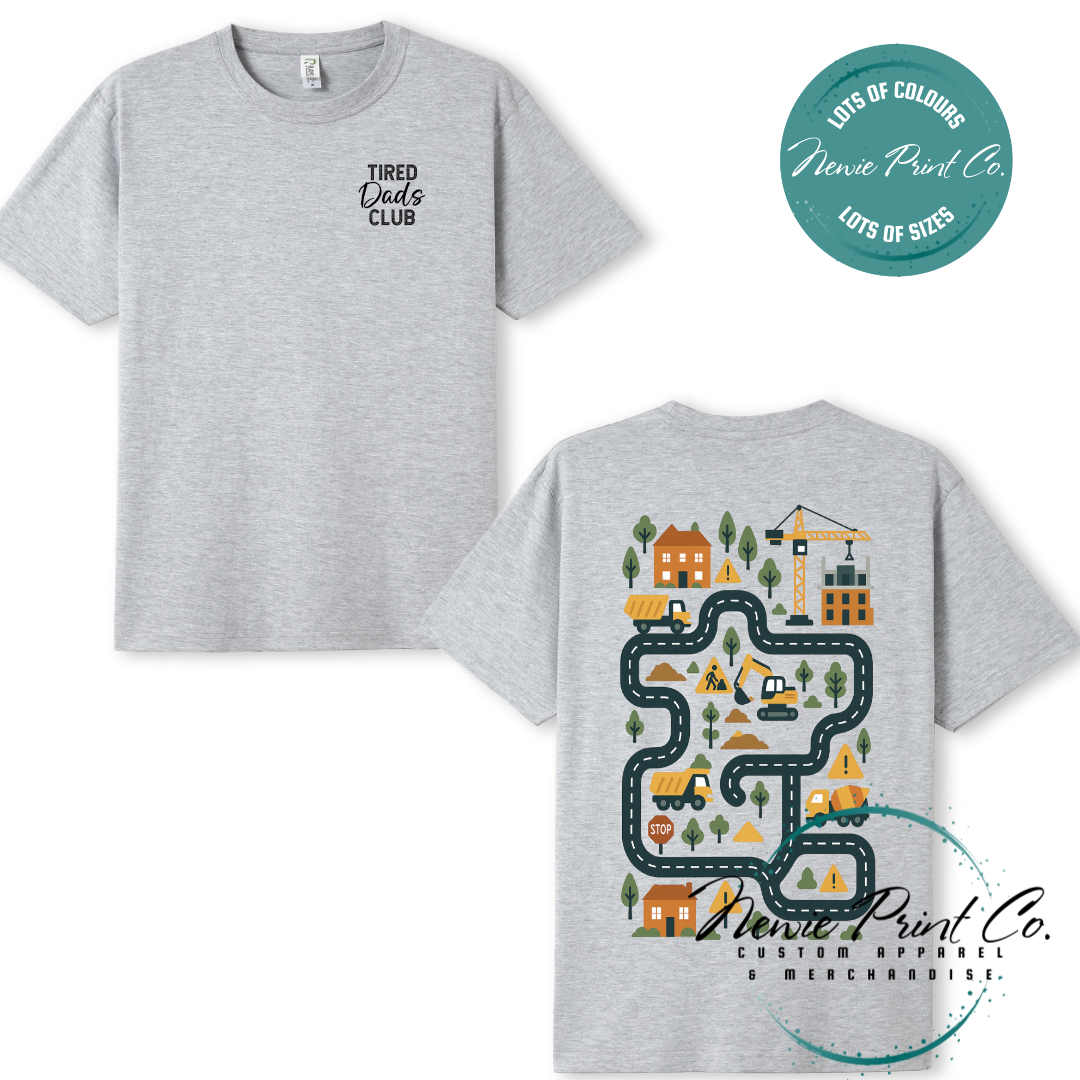 Tired Dads Club black t-shirt featuring front text and a vibrant construction-themed illustration on the back with houses, vehicles, and winding roads. Available in multiple t-shirt colors and various jumper options. Designed and printed by Newie Print Co.