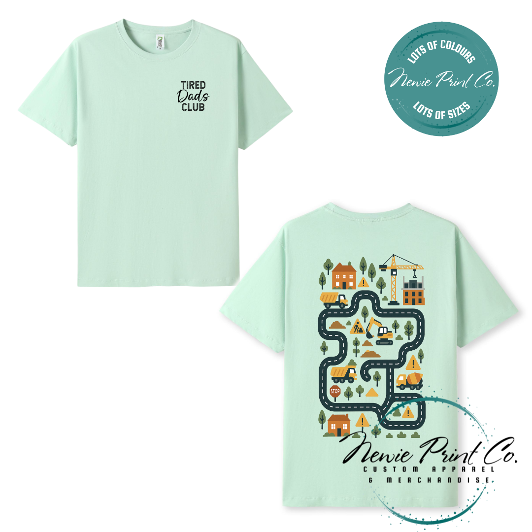 Tired Dads Club black t-shirt featuring front text and a vibrant construction-themed illustration on the back with houses, vehicles, and winding roads. Available in multiple t-shirt colors and various jumper options. Designed and printed by Newie Print Co.