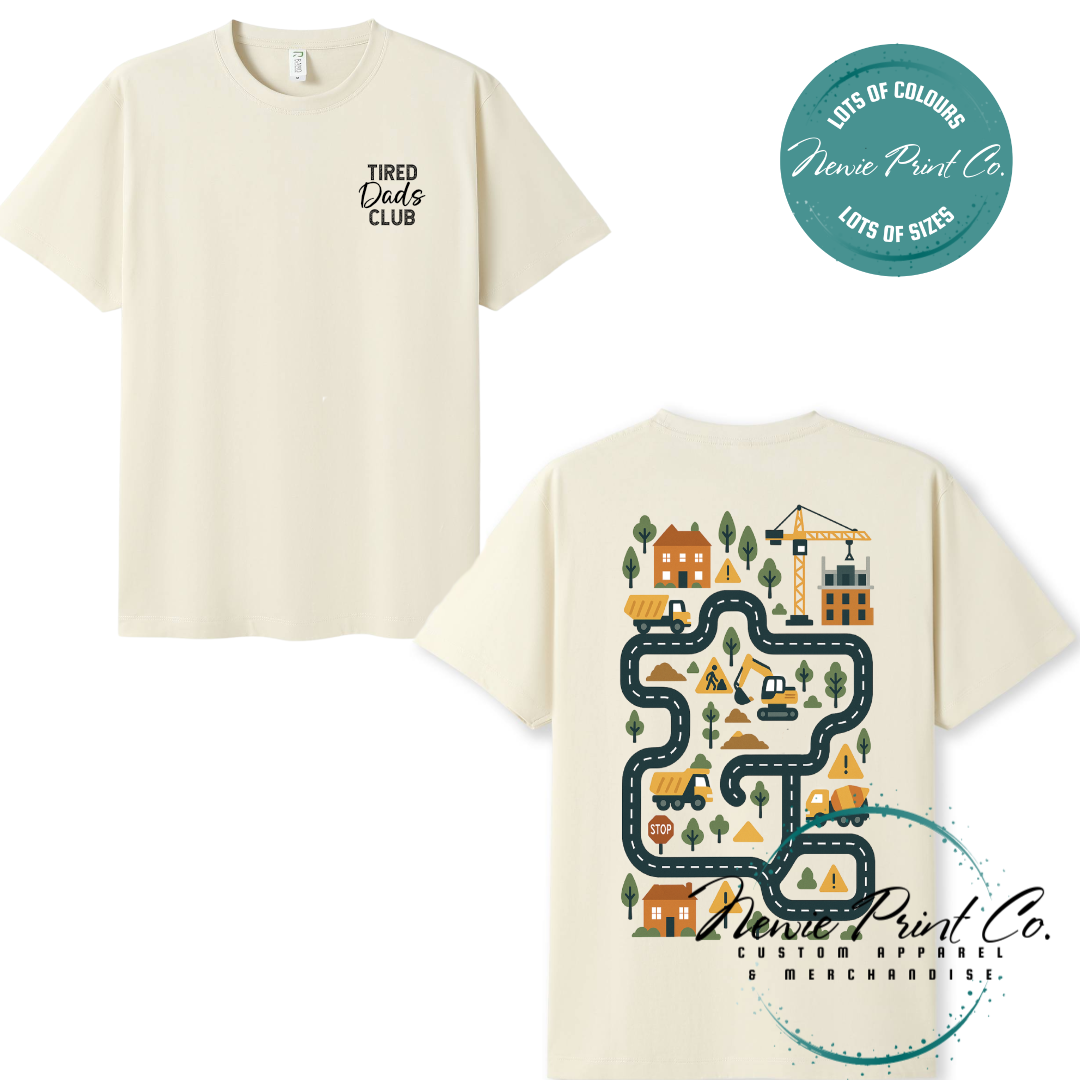 Tired Dads Club black t-shirt featuring front text and a vibrant construction-themed illustration on the back with houses, vehicles, and winding roads. Available in multiple t-shirt colors and various jumper options. Designed and printed by Newie Print Co.