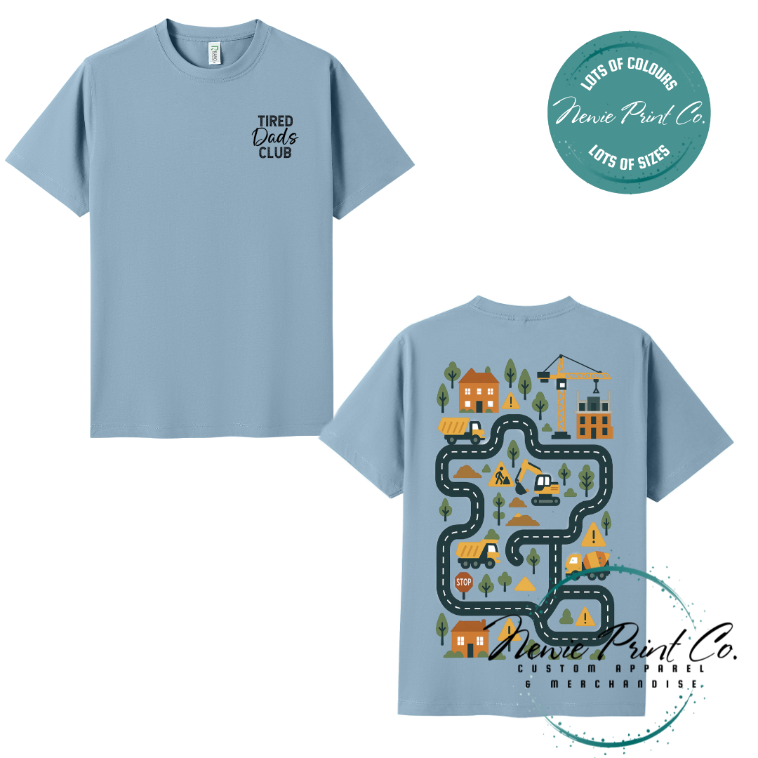 Tired Dads Club black t-shirt featuring front text and a vibrant construction-themed illustration on the back with houses, vehicles, and winding roads. Available in multiple t-shirt colors and various jumper options. Designed and printed by Newie Print Co.