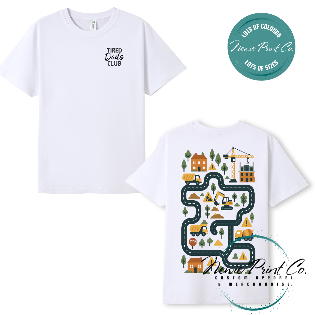 Tired Dads Club black t-shirt featuring front text and a vibrant construction-themed illustration on the back with houses, vehicles, and winding roads. Available in multiple t-shirt colors and various jumper options. Designed and printed by Newie Print Co.
