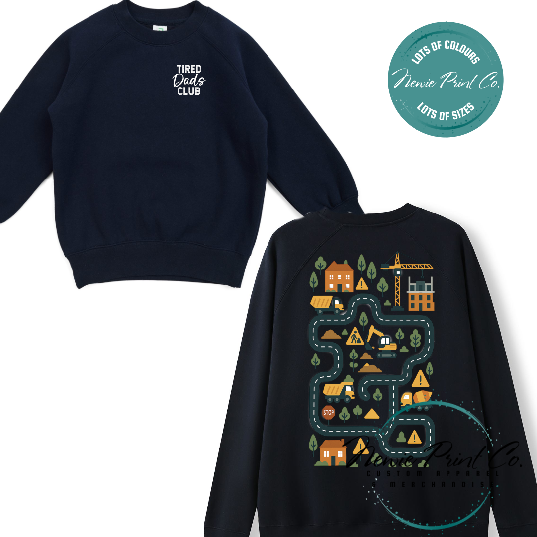 Tired Dads Club black t-shirt featuring front text and a vibrant construction-themed illustration on the back with houses, vehicles, and winding roads. Available in multiple t-shirt colors and various jumper options. Designed and printed by Newie Print Co.