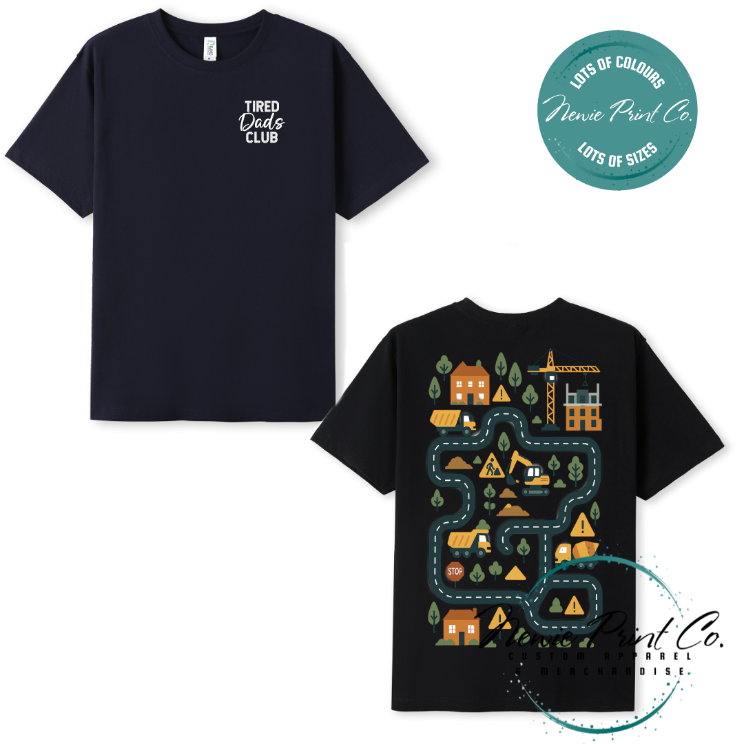 Tired Dads Club black t-shirt featuring front text and a vibrant construction-themed illustration on the back with houses, vehicles, and winding roads. Available in multiple t-shirt colors and various jumper options. Designed and printed by Newie Print Co.