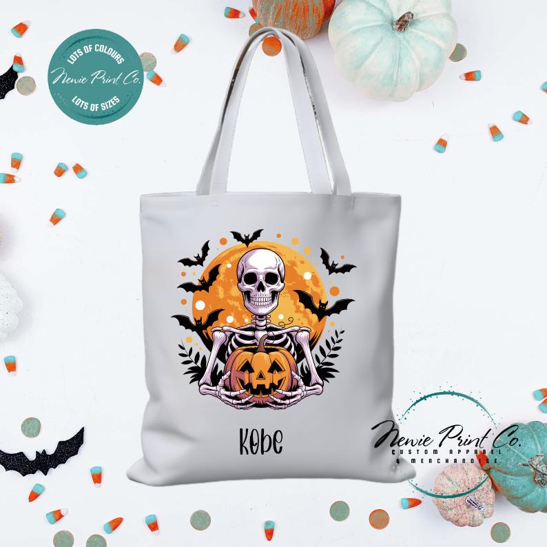 Custom Halloween tote bag with a skeleton holding a pumpkin and a big orange moon and bats behind him, optional personalised name printed on the front, perfect for trick-or-treating or spooky season gifts printed in Australia.