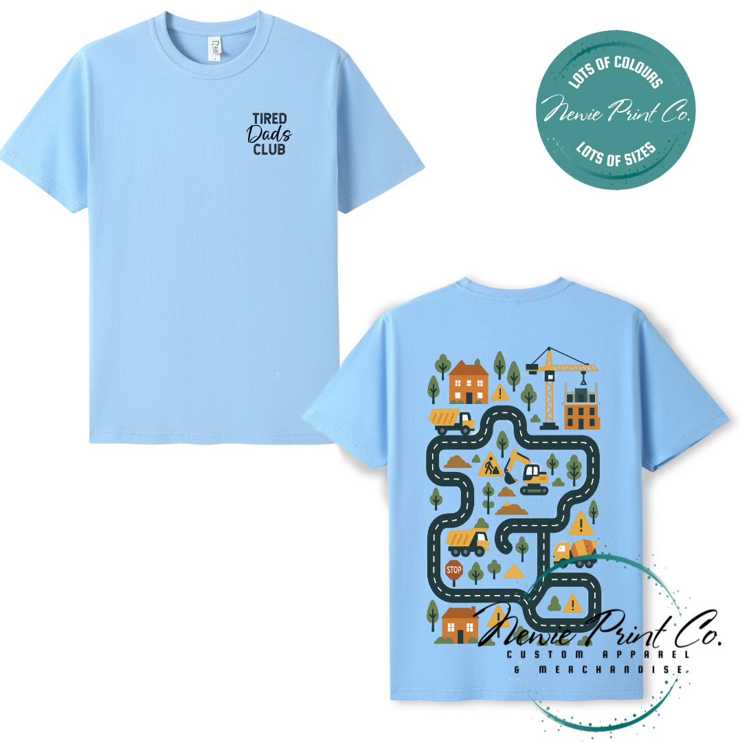 Tired Dads Club black t-shirt featuring front text and a vibrant construction-themed illustration on the back with houses, vehicles, and winding roads. Available in multiple t-shirt colors and various jumper options. Designed and printed by Newie Print Co.