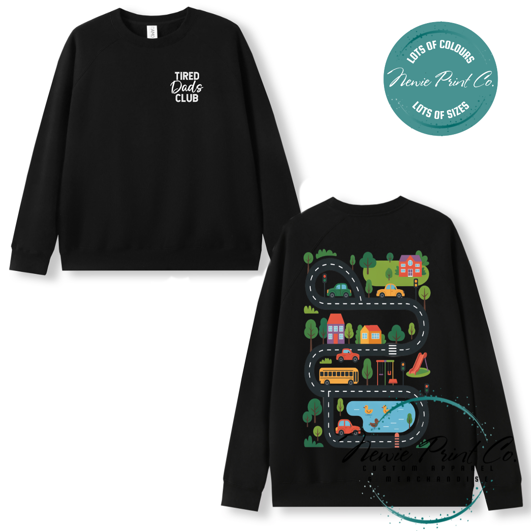 Tired Dads Club black t-shirt featuring front text and a vibrant town-themed illustration on the back with houses, vehicles, and winding roads. Available in multiple t-shirt colors and various jumper options. Designed and printed by Newie Print Co.