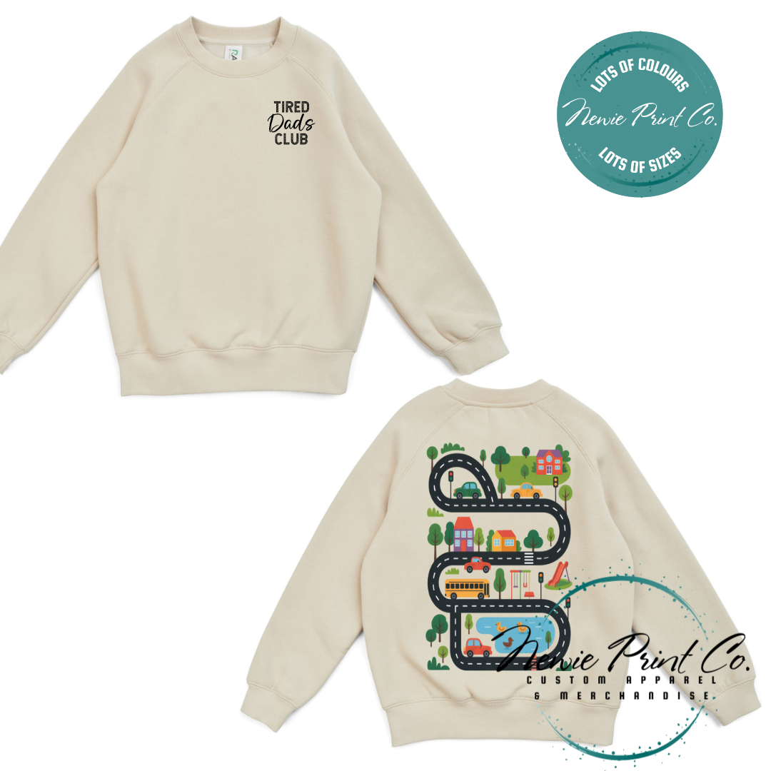 Tired Dads Club black t-shirt featuring front text and a vibrant town-themed illustration on the back with houses, vehicles, and winding roads. Available in multiple t-shirt colors and various jumper options. Designed and printed by Newie Print Co.