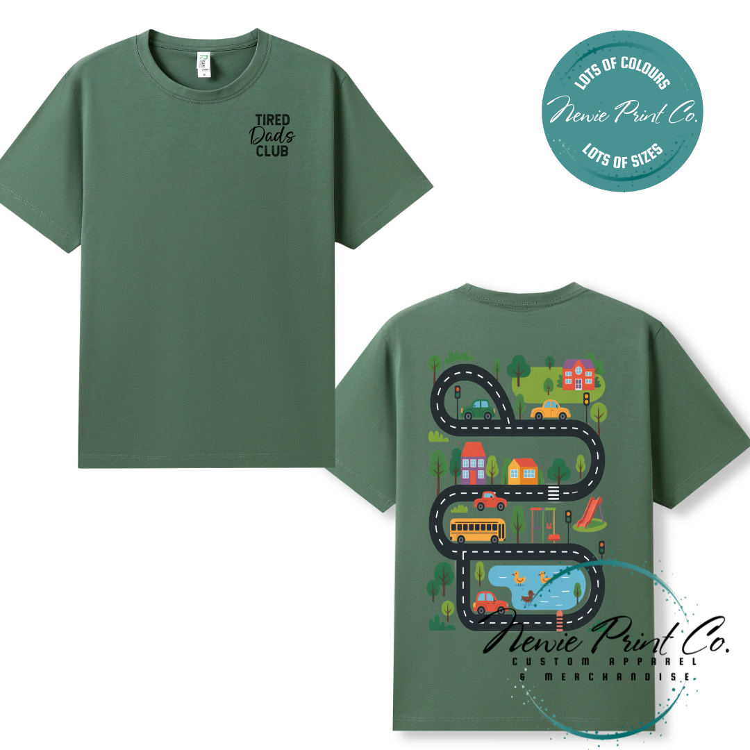 Tired Dads Club black t-shirt featuring front text and a vibrant town-themed illustration on the back with houses, vehicles, and winding roads. Available in multiple t-shirt colors and various jumper options. Designed and printed by Newie Print Co.