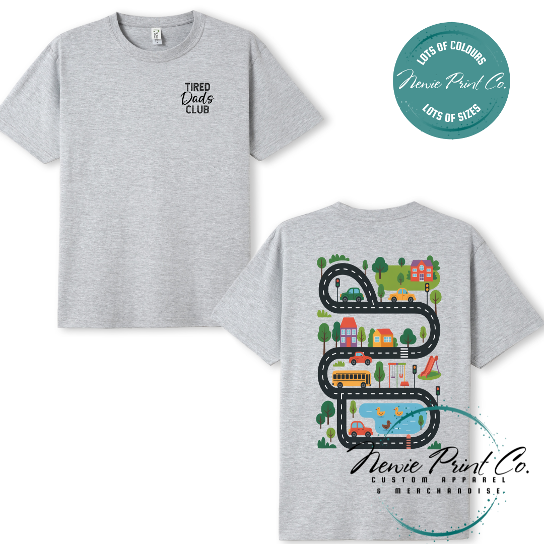 Tired Dads Club black t-shirt featuring front text and a vibrant town-themed illustration on the back with houses, vehicles, and winding roads. Available in multiple t-shirt colors and various jumper options. Designed and printed by Newie Print Co.