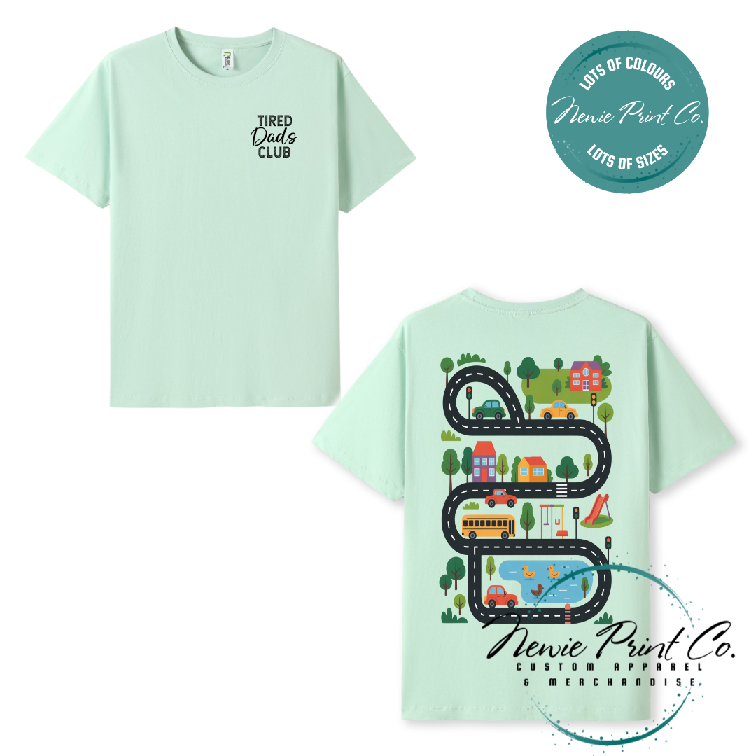 Tired Dads Club black t-shirt featuring front text and a vibrant town-themed illustration on the back with houses, vehicles, and winding roads. Available in multiple t-shirt colors and various jumper options. Designed and printed by Newie Print Co.
