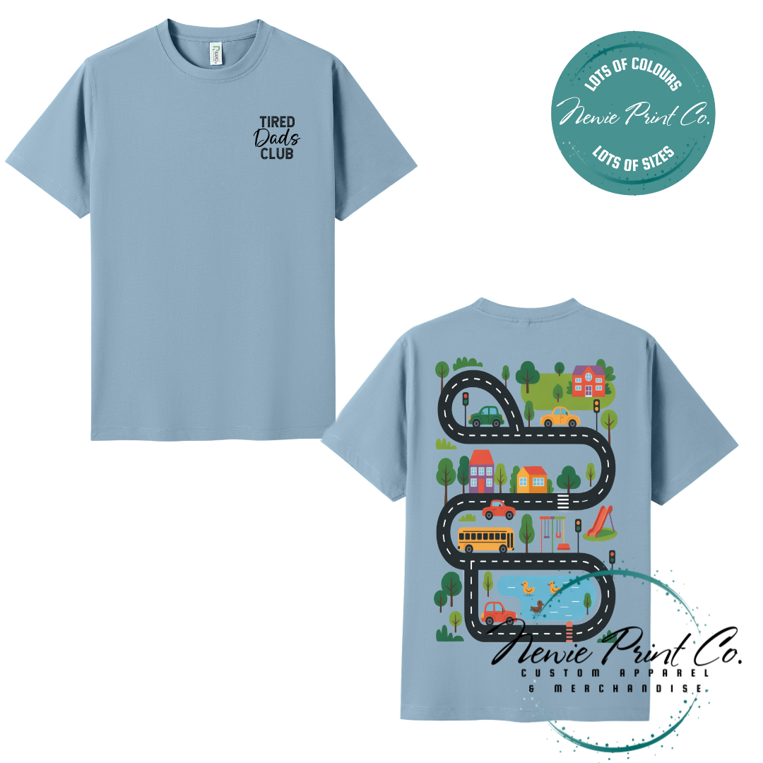 Tired Dads Club black t-shirt featuring front text and a vibrant town-themed illustration on the back with houses, vehicles, and winding roads. Available in multiple t-shirt colors and various jumper options. Designed and printed by Newie Print Co.