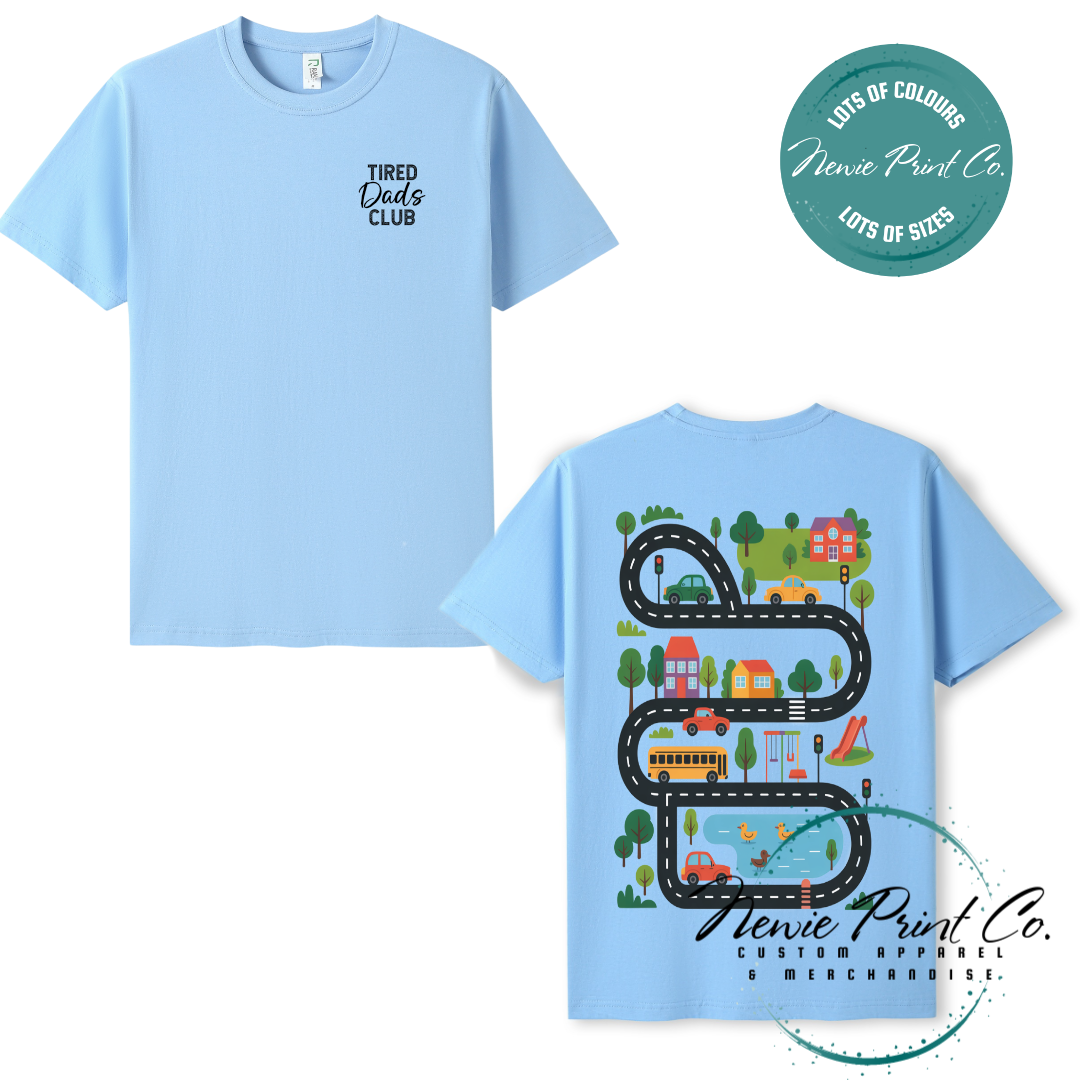 Tired Dads Club black t-shirt featuring front text and a vibrant town-themed illustration on the back with houses, vehicles, and winding roads. Available in multiple t-shirt colors and various jumper options. Designed and printed by Newie Print Co.