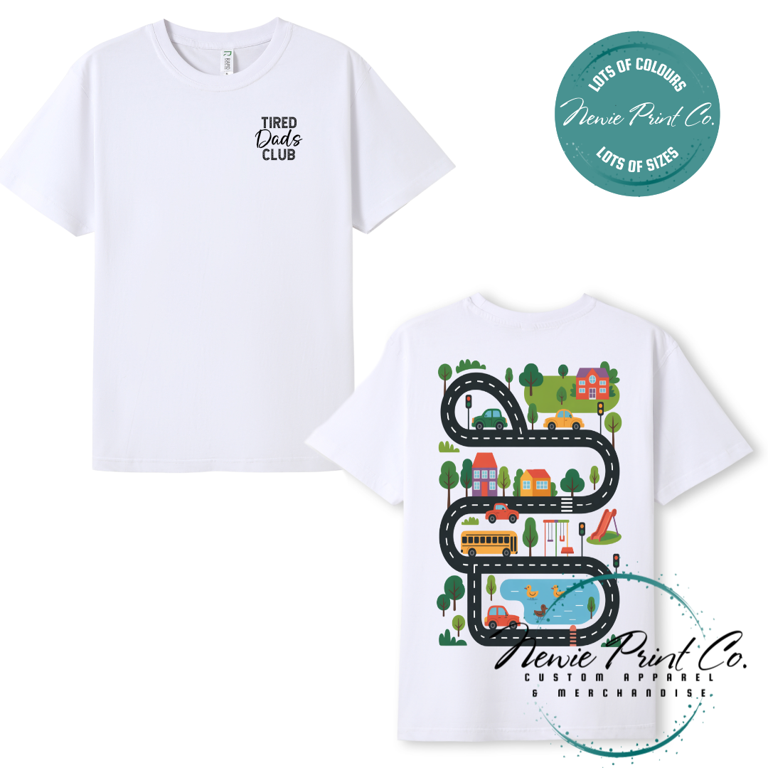 Tired Dads Club black t-shirt featuring front text and a vibrant town-themed illustration on the back with houses, vehicles, and winding roads. Available in multiple t-shirt colors and various jumper options. Designed and printed by Newie Print Co.