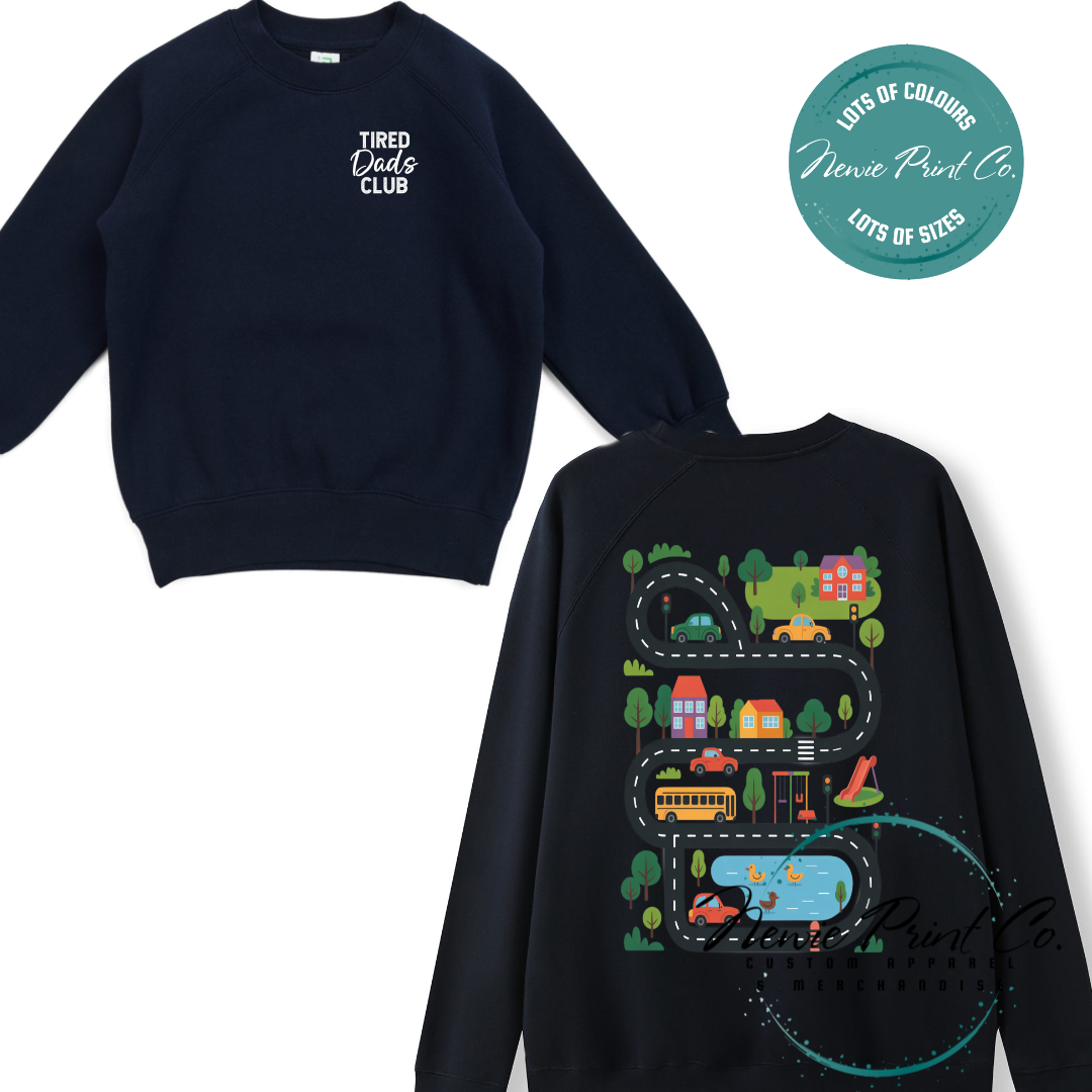 Tired Dads Club black t-shirt featuring front text and a vibrant town-themed illustration on the back with houses, vehicles, and winding roads. Available in multiple t-shirt colors and various jumper options. Designed and printed by Newie Print Co.
