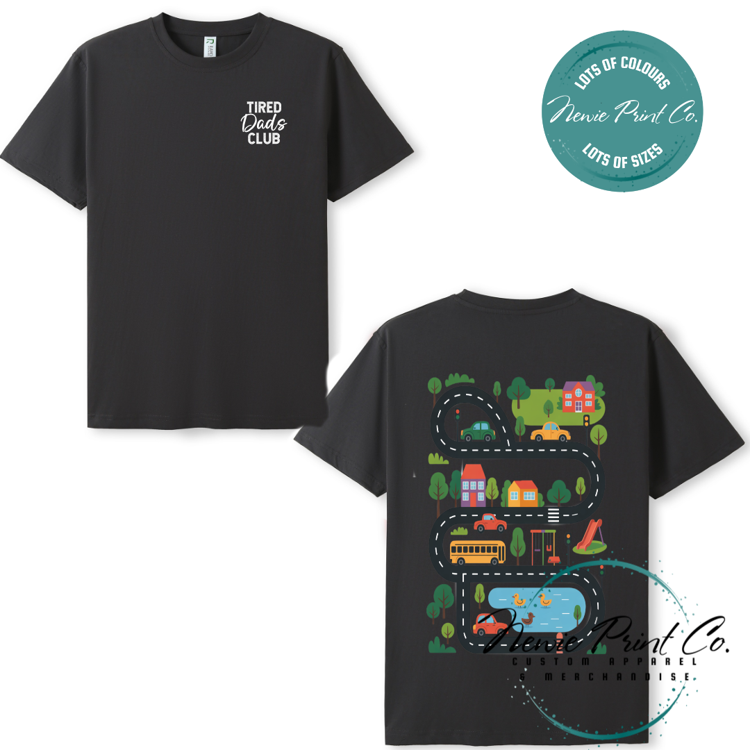Tired Dads Club black t-shirt featuring front text and a vibrant town-themed illustration on the back with houses, vehicles, and winding roads. Available in multiple t-shirt colors and various jumper options. Designed and printed by Newie Print Co.