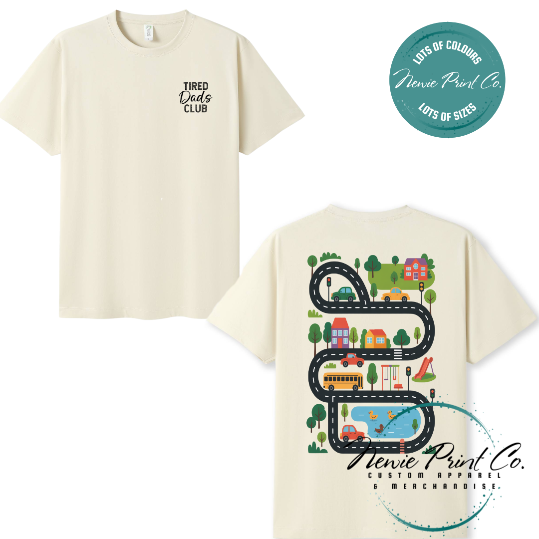 Tired Dads Club black t-shirt featuring front text and a vibrant town-themed illustration on the back with houses, vehicles, and winding roads. Available in multiple t-shirt colors and various jumper options. Designed and printed by Newie Print Co.