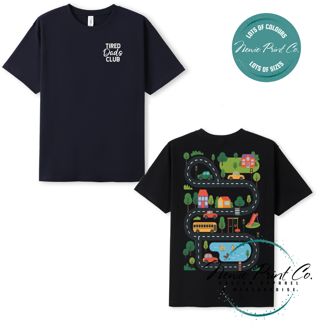Tired Dads Club black t-shirt featuring front text and a vibrant town-themed illustration on the back with houses, vehicles, and winding roads. Available in multiple t-shirt colors and various jumper options. Designed and printed by Newie Print Co.