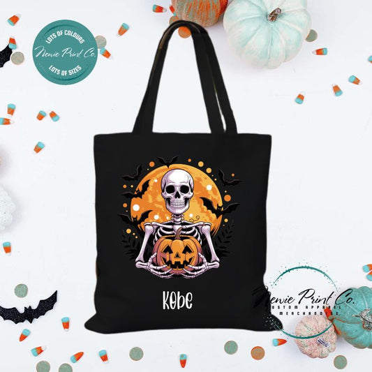 Custom Halloween tote bag with a skeleton holding a pumpkin and a big orange moon and bats behind him, optional personalised name printed on the front, perfect for trick-or-treating or spooky season gifts printed in Australia by Newie Print Co.